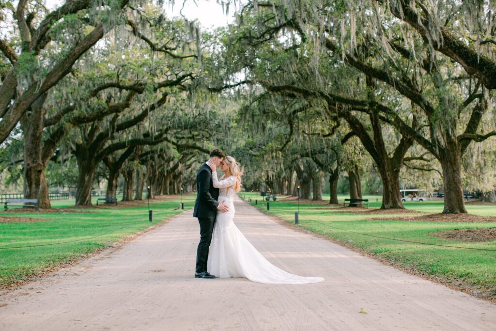a Groom in black Tuxedo and Bride kissing on a path with trees with Spanish Moss hanging at Boone Hall Wedding Venue Charleston, South Carolina