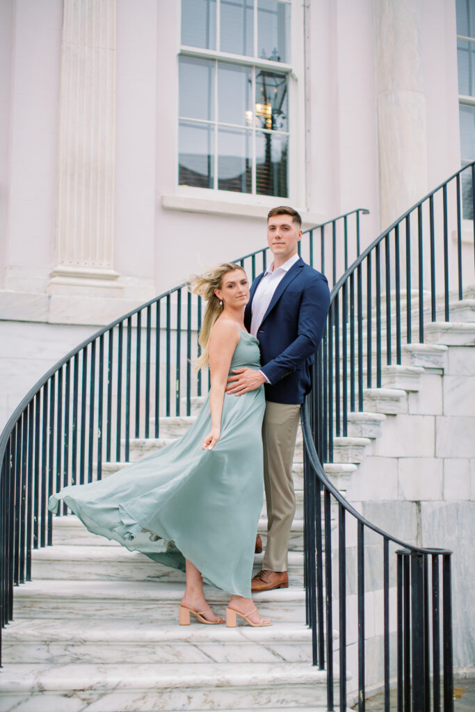a man with khaki pants and blue sports jacket and woman with sage green long dress posing on stairs at the charleston courthouse on broad street