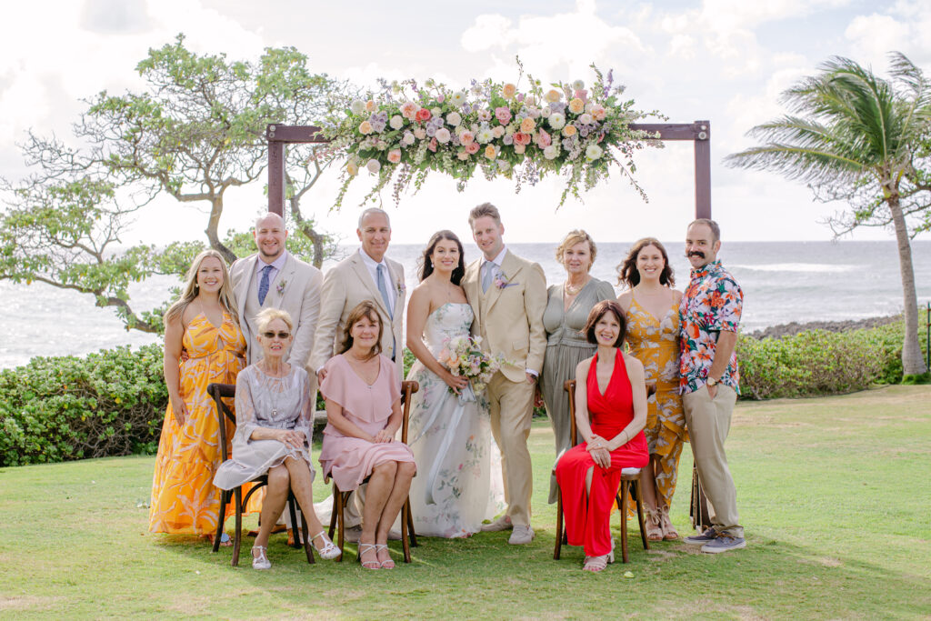 Family group picture at Turtle Bay Resort Destination Wedding showing the bride and groom with family members An Arbor of pink and white florals with the pacific ocean in the background. The bride wears a Monique Lhuillier gown with floral design.