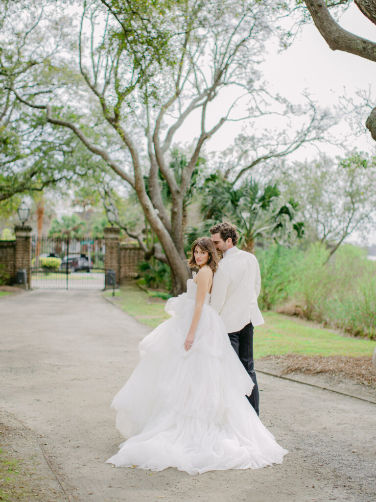Bride and Groom walking  along the Oak Lined pathway at Lowndes Grove in Charleston, South Carolina. the bride is in a strapless tule layered dress and the groom is wearing a white tuxedo jacket and black pants.
