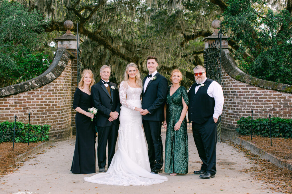 Family Portrait at Boone Hall Plantation in front of the grand entrance iron gate. Mother of the bride is wearing a black eveing gown and Father is wearing a tuxedo with black bow tie. Mother of the groom is wearing a  green sequence dress and Father of the groom is wearing black vest with bow tie.
