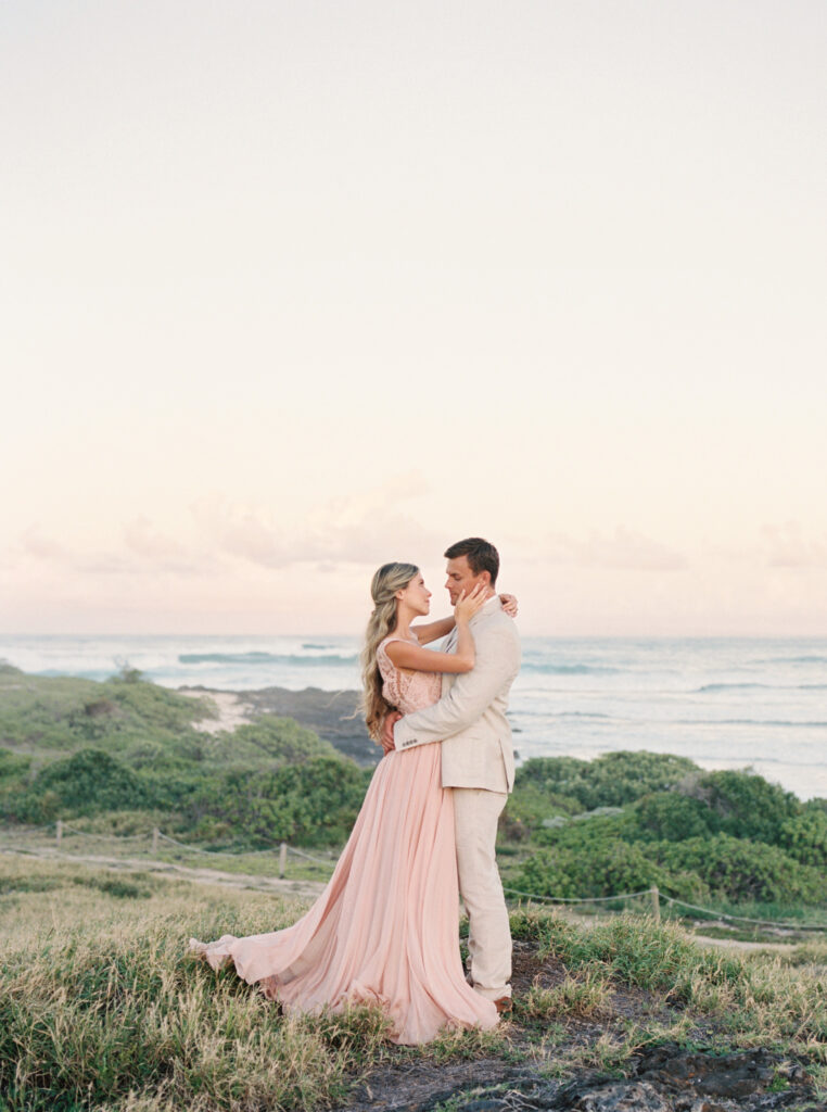 a groom and bride in a pink dress on the beach in Hawaii at sunset